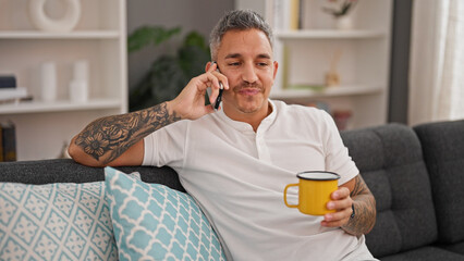 Wall Mural - Young hispanic man talking on smartphone drinking coffee at home