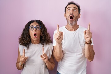 Canvas Print - Middle age hispanic couple together over pink background amazed and surprised looking up and pointing with fingers and raised arms.