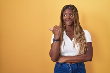 Sticker - African american woman with braided hair standing over yellow background smiling with happy face looking and pointing to the side with thumb up.