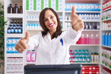 Poster - Middle age brunette woman working at pharmacy drugstore approving doing positive gesture with hand, thumbs up smiling and happy for success. winner gesture.