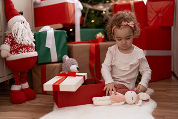 Canvas Print - Adorable caucasian girl unpacking gift sitting on floor by christmas tree at home