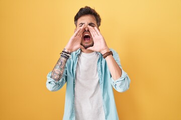 Canvas Print - Young hispanic man with tattoos standing over yellow background shouting angry out loud with hands over mouth