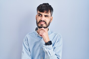 Wall Mural - Young hispanic man with beard standing over blue background thinking worried about a question, concerned and nervous with hand on chin