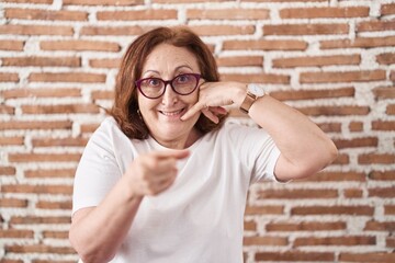 Poster - Senior woman with glasses standing over bricks wall smiling doing talking on the telephone gesture and pointing to you. call me.