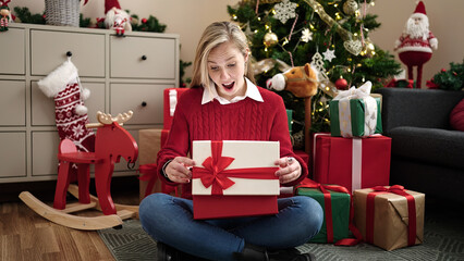 Wall Mural - Young blonde woman unpacking gift sitting on floor by christmas tree at home