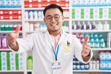 Poster - Chinese young man working at pharmacy drugstore looking at the camera smiling with open arms for hug. cheerful expression embracing happiness.