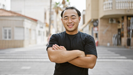 Sticker - Cheerful young chinese man, radiating confidence and joy, spotted smiling and standing with crossed arms gesture on a bustling urban street