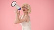 Woman in character of speaking into megaphone. Woman with colorful makeup, wig and white dress in studio on pink background. Sale, Black Friday. Copy space.