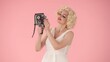 © kinomaster - Woman taking selfies using old vintage video camera. Woman in look like poses for camera on pink background in studio.