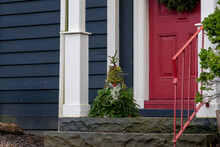 A Christmas Gnome Decoration On The Step Of A Blue House With A Vibrant Red Door, White Columns, And A Red Rail.The Character Is Made Of Green Spruce Tree Boughs, Moss, And A Red Ornament For His Nose