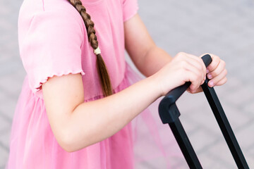 Close-up of a little girl with a suitcase. Children's hands hold the handle of a travel bag. A child holds a large black suitcase by the handle at a train station. Tourism, travel concept, vacation