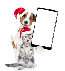 Wall Mural - Happy Jack russell terrier puppy and funny cute kitten wearing santa hats standing together showing big smartphone with white blank screen in it paw. isolated on white background