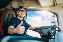 Handsome experienced male truck driver with a hat sitting and driving his truck. He is looking at camera and showing thumb up. Professional transportation and truck drivers concept.
