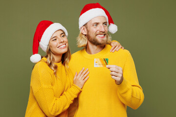 Canvas Print - Merry young couple two friends man woman in sweater Santa hat posing use mobile cell phone credit bank card pay look aside isolated on plain green background Happy New Year Christmas holiday concept