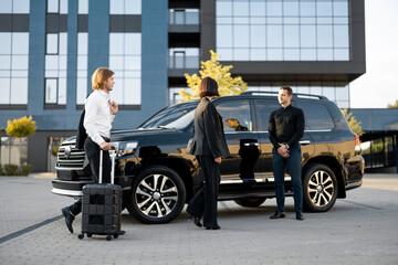 Wall Mural - Businessman and businesswoman walk with a suitcase to a luxury black car during a business trip. Male chauffeur waiting near vehicle. Concept of transportation and business travel