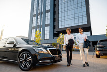 Two Businessmen Walk To A Luxury Black Car Near Hotel Or Office Building On Sunset. Concept Of Transportation And Business Lifestyle