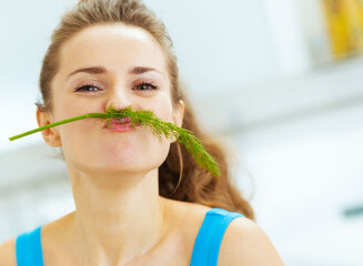 Wall Mural - Funny young woman using dill as mustache in kitchen