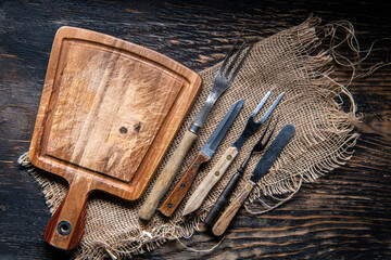 Wall Mural - Wood cutting board with a piece of burlap, knives and forks on wooden table with copy space, top view