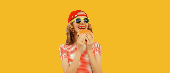 Wall Mural - Portrait of stylish happy smiling young woman eating burger fast food on yellow studio background