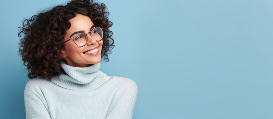 Canvas Print - Horizontal photograph of a self embracing and joyful woman who appears optimistic but shy donning transparent glasses and a casual turtleneck standing alone against a blue background with am