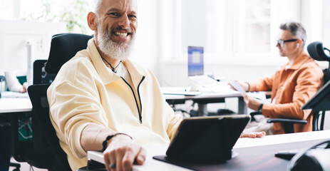 Wall Mural - Cheerful male entrepreneur with modern touch pad device laughing during working time in office interior, portrait of joyful Caucasian employee with digital tablet posing at table desktop