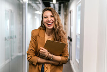 Happy Businesswoman With Clipboard Standing In Corridor At Office