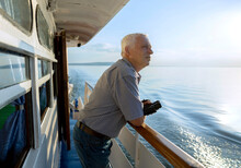 Senior Man With Binoculars In Hand Looking At Sea From Ship On Sunny Day