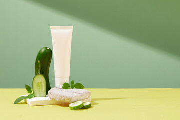 Wall Mural - Front view of a white plastic tube displayed on bricks podiums with fresh cucumber slices and mint leaves on green and yellow background. Unlabeled bottle for design packaging and copy space