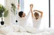 Back view of caucasian couple in casual attire stretching their limbs after night sleep while in bed of bright bedroom. Relaxed family man and woman starting new sunny day in modern flat.