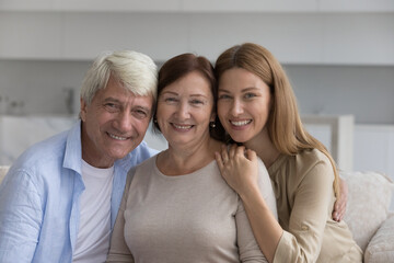 Wall Mural - Older man posing for family portrait, hugging mature senior wife and adult daughter, looking at camera, smiling. Happy woman visiting couple of elder parents