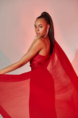 Wall Mural - charming african american woman in red dress posing with shawl on grey backdrop with lighting