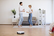 © sofiko14 - Young caucasian lady dancing with male partner while robotic vacuum running floor smoothly. Homeowners using electrical appliance for household chores while freeing up time for mother-daughter party.
