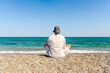 A man sits on the beach in the lotus position and looks at the sea. Meditating man in summer clothes on the beach.