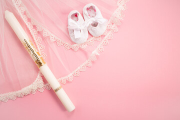 Christening background with baptism baby shoes, dress and candle on pink background.