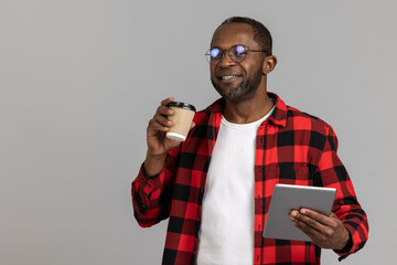 Cheerful black bearded man wearing red checkered shirt using tablet and drinking takeaway coffee