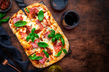 Wall Mural - Rectangular roman pizza with smoked ham, mozzarella cheese, sun dried tomatoes, tomato sauce and green basil leaves on rustic wooden table background, top view