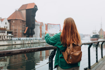 Wall Mural - Attractive young female tourist is exploring new city. Redhead girl with backpack holding a paper map on city street in Gdansk. Traveling Europe in autumn. Famous Zuraw crane, Motlawa river