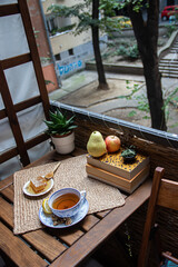 Wall Mural - Morning setup on wooden table at balcony, books to read, cup of natural tea, teapot, organic honey from farm, fresh green tea leaves and organic fruits, in background nice view on small city park 