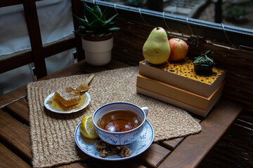 Wall Mural - Morning setup on wooden table at balcony, books to read, cup of natural tea, teapot, organic honey from farm, fresh green tea leaves and organic fruits, in background nice view on small city park 