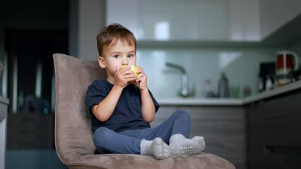 Wall Mural - Serious calm kid sits on the chair with his feet up. Cute baby boy eating the apple. Blurred backdrop.