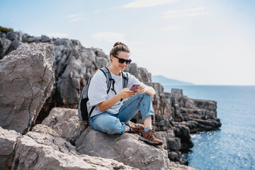 Wall Mural - Woman traveler hiker relaxing on a rocky seashore with a smartphone in her hands. Summer travel and adventure concept
