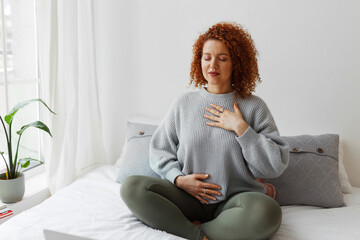 Fototapete - Portrait of plus-size curly redhead female in casual clothes doing pranayama technique sitting on bed next to window with closed eyes, putting hands on chest, breathing slowly and deeply