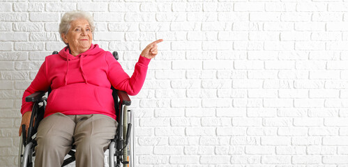 Poster - Senior woman in wheelchair pointing at something near white brick wall with space for text