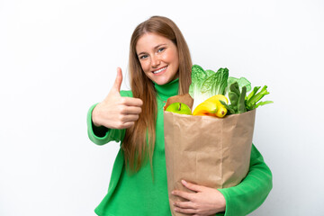 Wall Mural - Young caucasian woman holding a grocery shopping bag isolated on white background with thumbs up because something good has happened