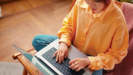 Wall Mural - Portrait of a young bright Asian woman with pink streaks of hair sitting in a chair with a laptop and taking online training or working in the living room.