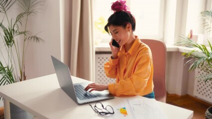 Wall Mural - Asian young girl with bright pink locks of hair sitting at a table with a laptop at home in the living room, talking on the phone, working or taking online training.