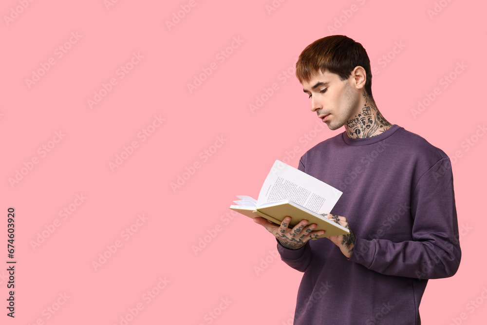 Young tattooed man reading book on pink background