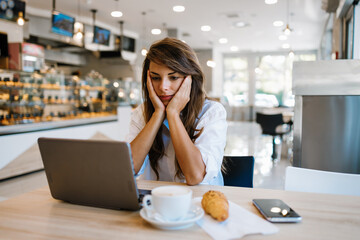 Beautiful and happy young woman sitting and eating delicious rolls in bakery or fast food while she using laptop computer. She is serious and tired.