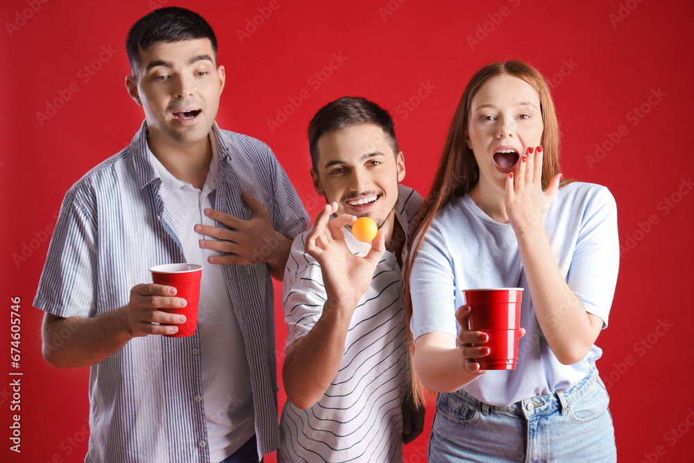 Young friends with cups and ball for beer pong on red background