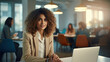 © Sasint - Beautiful Middle Eastern Manager Sitting at a Desk in Creative Office. Young Stylish Female with Curly Hair Using Laptop Computer in Marketing Agency. Colleagues Working in the Background